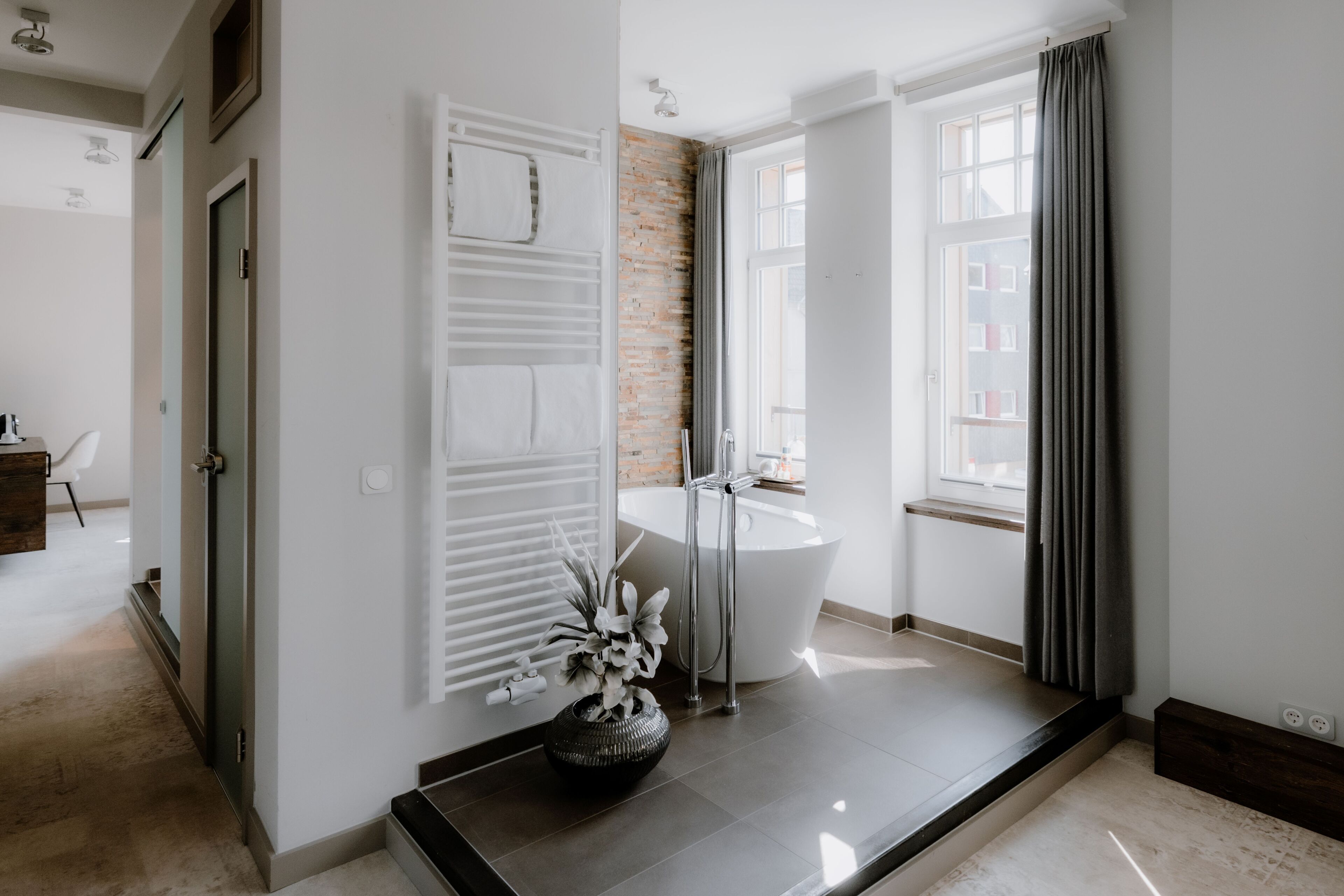 A modern bathroom with a bathtub, a shower, and a sink. There is a door leading to another room, and a window with curtains.