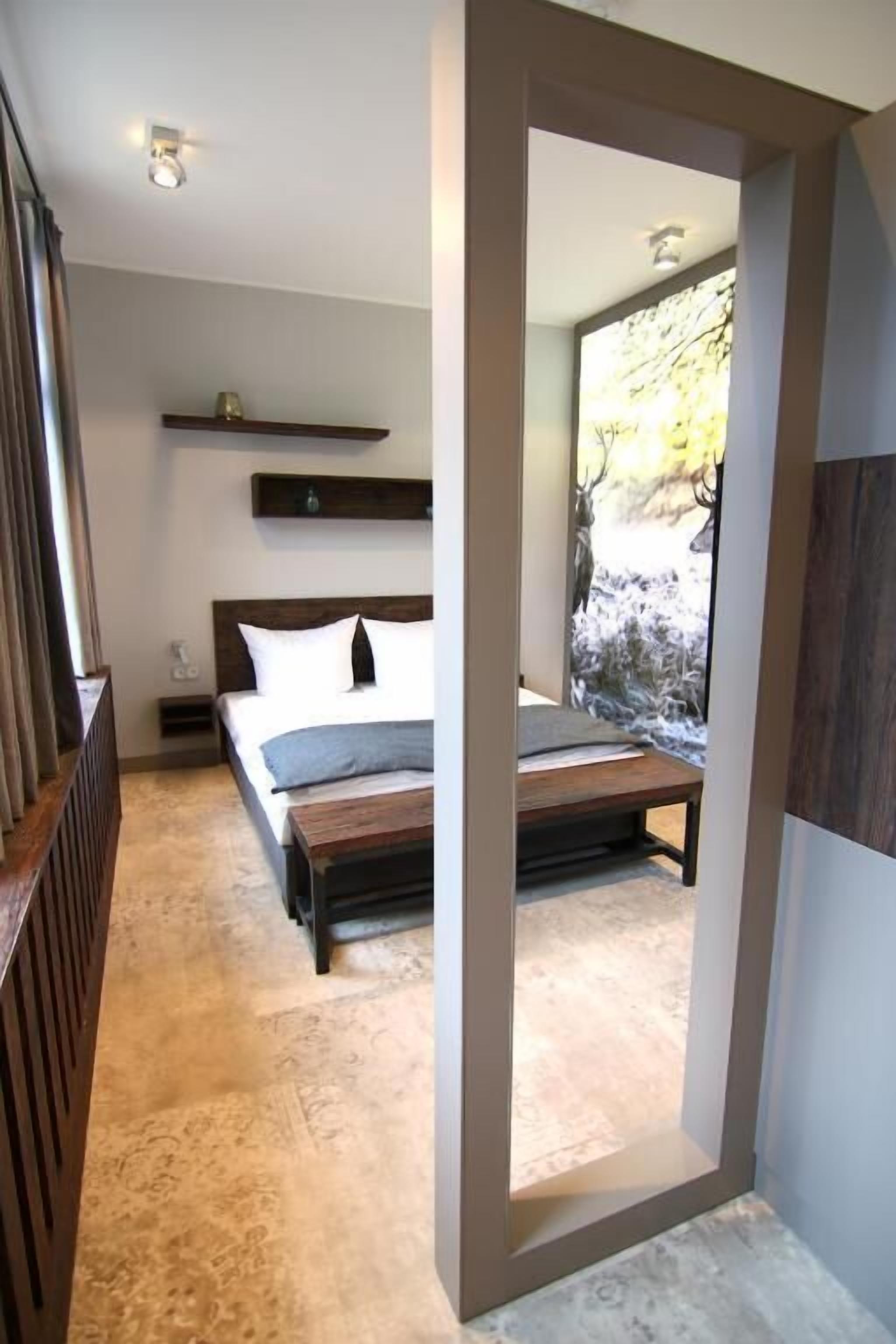 Un dormitorio con una cama, una mesita de noche y un espejo que refleja una habitación con ventanal.