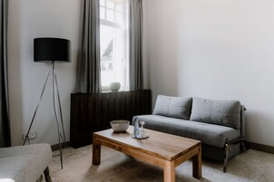 Una sala de estar con mesa de centro de madera, sofá gris, lámpara de pie y una ventana con cortinas.
