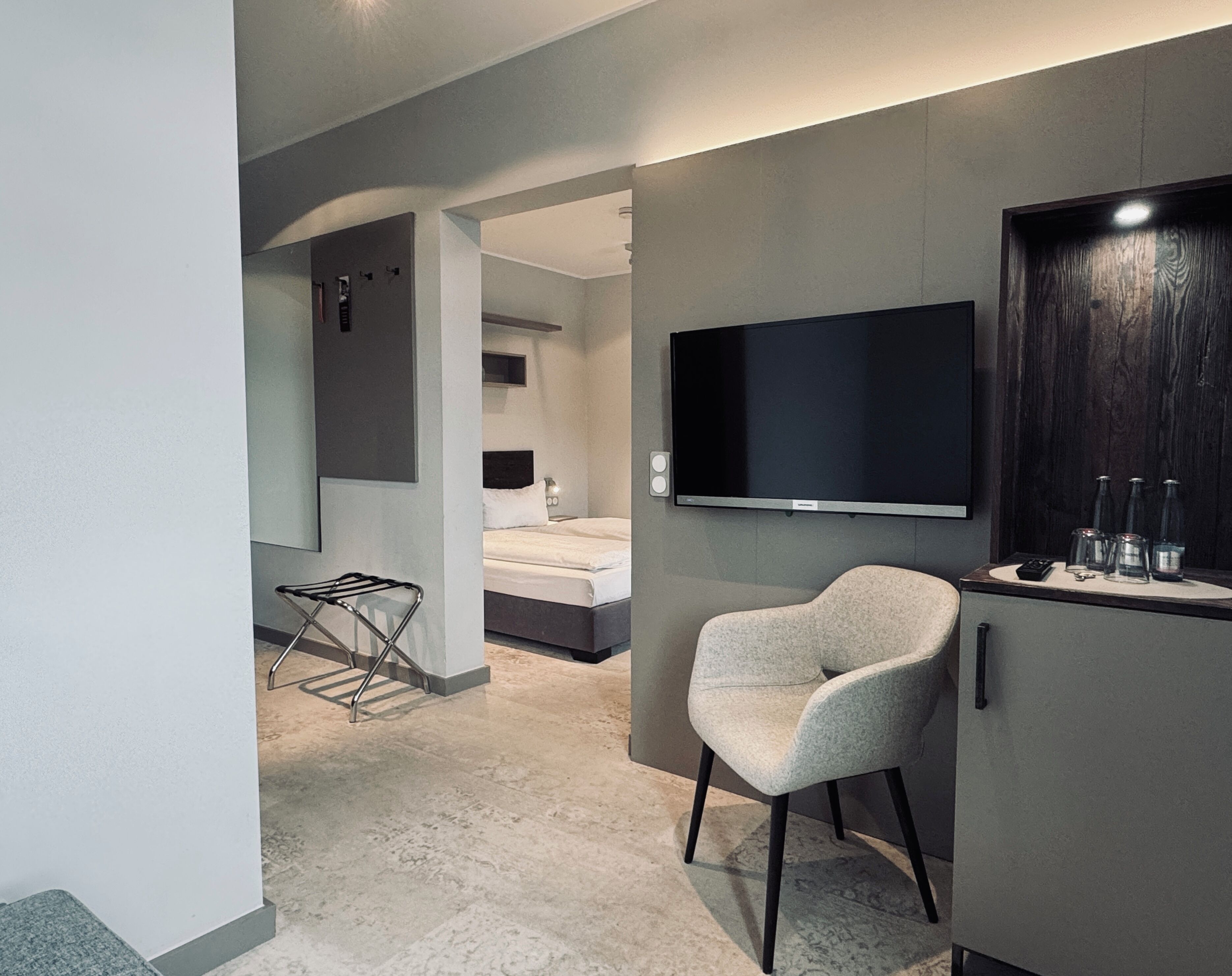 Habitación de hotel moderna con cama, zona de estar con una silla, televisor de pantalla plana montado en la pared y minibar.
