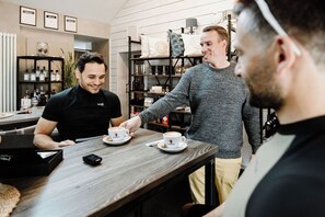 Un café con un barista sirviendo café a los clientes. Se observan estantes con diversos artículos y una barra con una computadora portátil y un teléfono móvil.