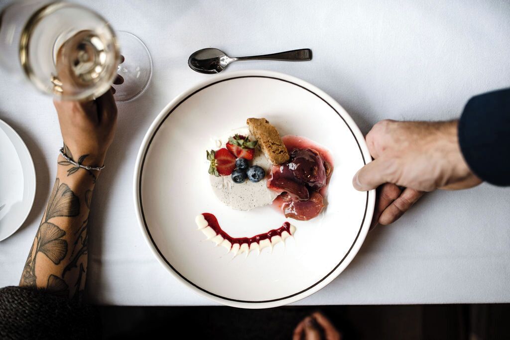 Eine Person hält einen Teller mit Dessert, einem Weinglas und einem Löffel auf einem Tisch.
