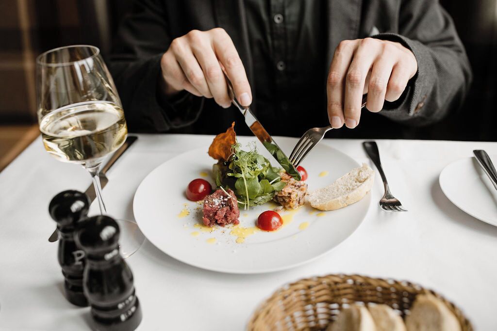 Una persona cenando en una mesa con un plato de comida, una copa de vino blanco y saleros y pimenteros.