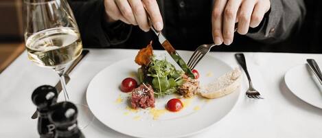 Una persona cenando en una mesa con un plato de comida, una copa de vino blanco y saleros y pimenteros.