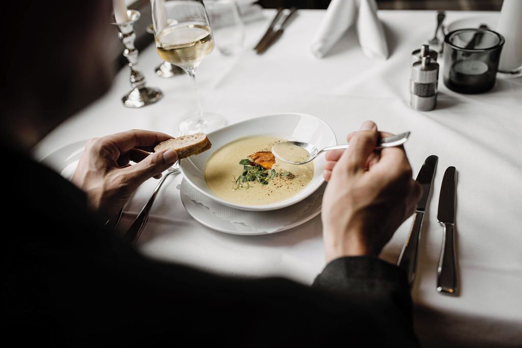 A person dining with a bowl of soup and bread, a glass of white wine, and cutlery on a table.