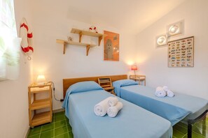 Ein Hotelzimmer mit zwei Betten, einem hölzernen Kopfteil, einem Regal mit Dekoration, einem Wandbild eines Geistes, einer Wandtafel mit Fischmotiven und einem grün gefliesten Boden.