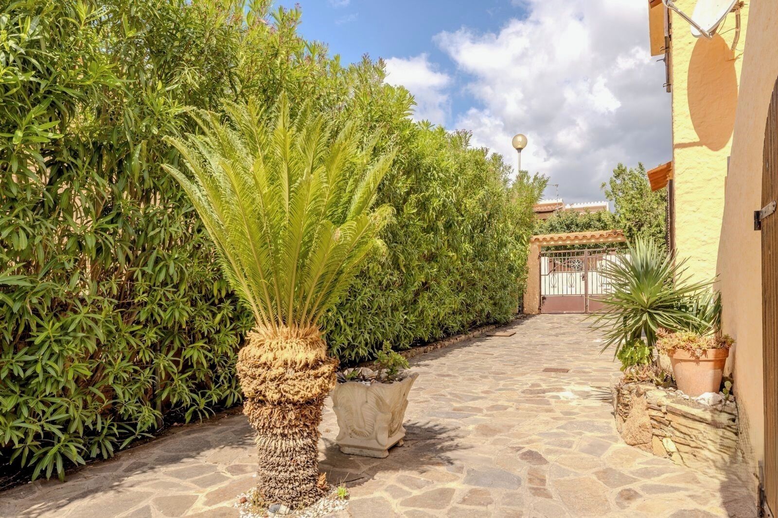 A stone-paved pathway leading to a gate, surrounded by lush greenery and a building with a yellow facade.