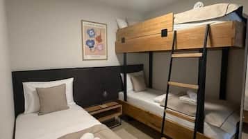 Quarto com beliche e uma cama de solteiro à esquerda, e o beliche à direita. Um criado-mudo com uma luminária fica entre as camas.