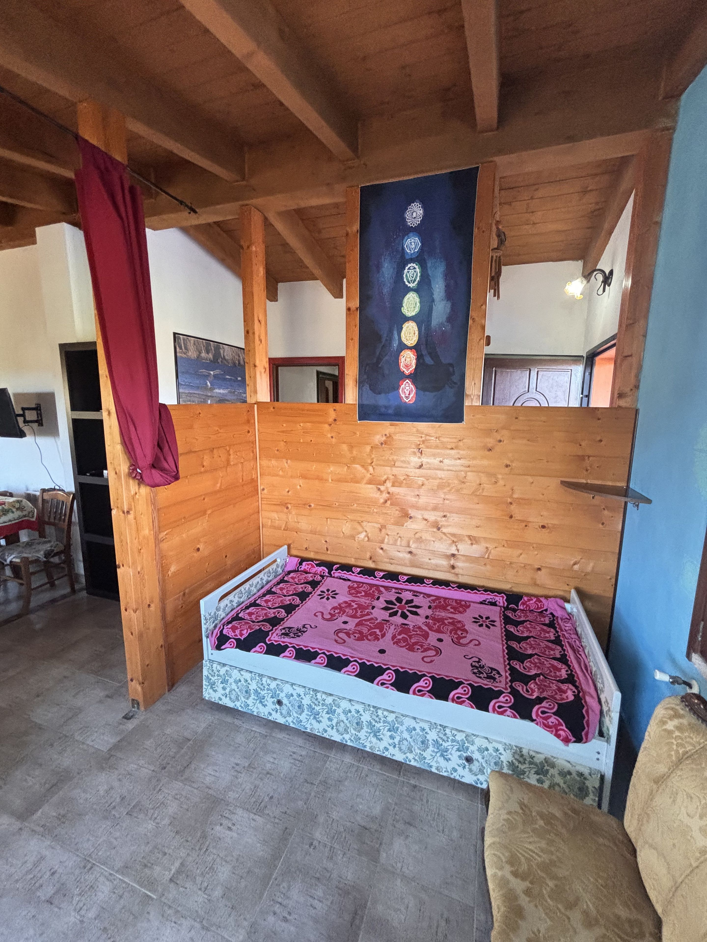 Ein Zimmer mit Holzboden, einem Bett mit gemustertem Überwurf, einem roten Vorhang und einer Wand mit einem Poster.