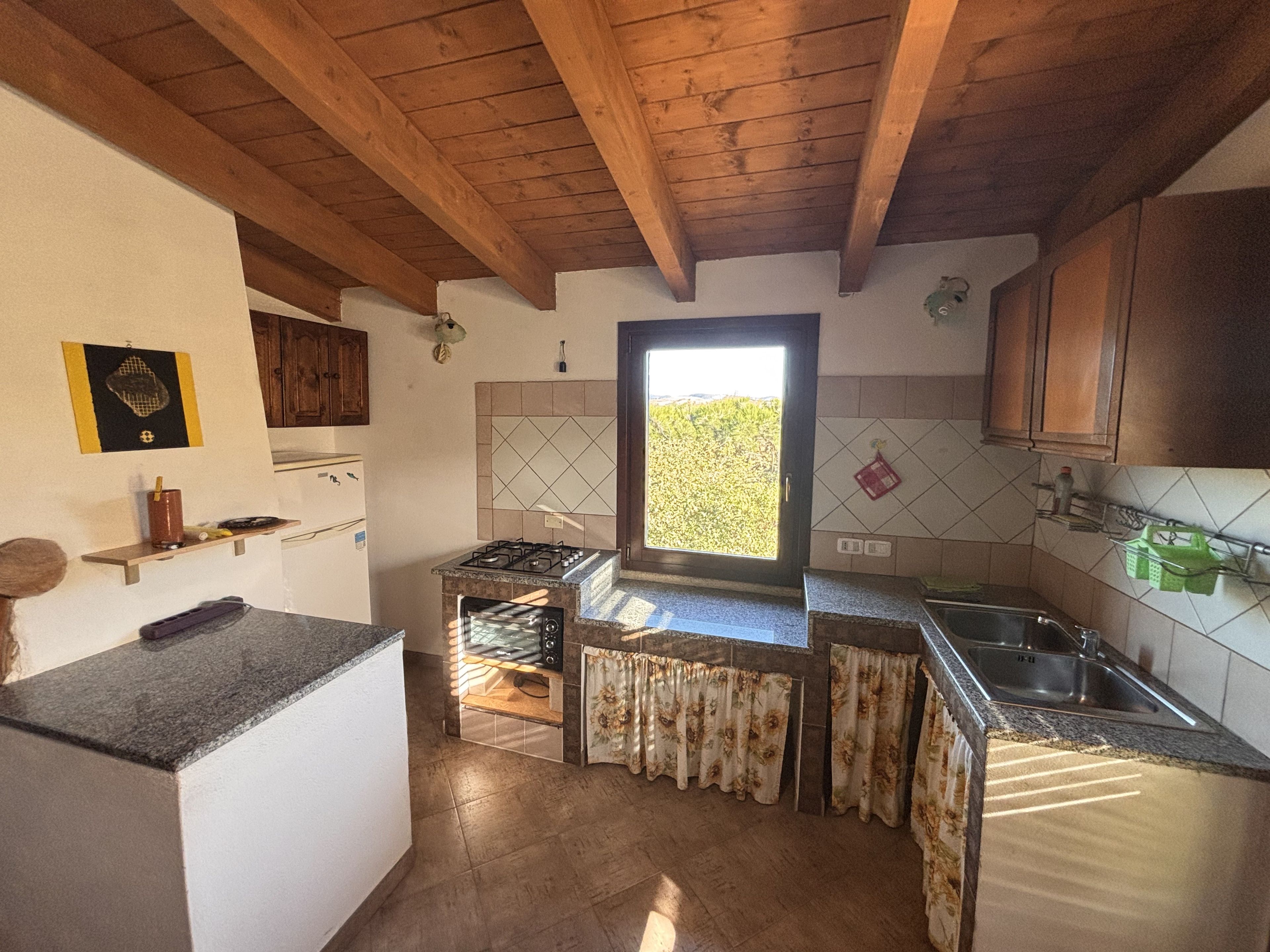 A compact kitchen with wooden beams, a gas stove, a built-in oven, and a window with a view of greenery.