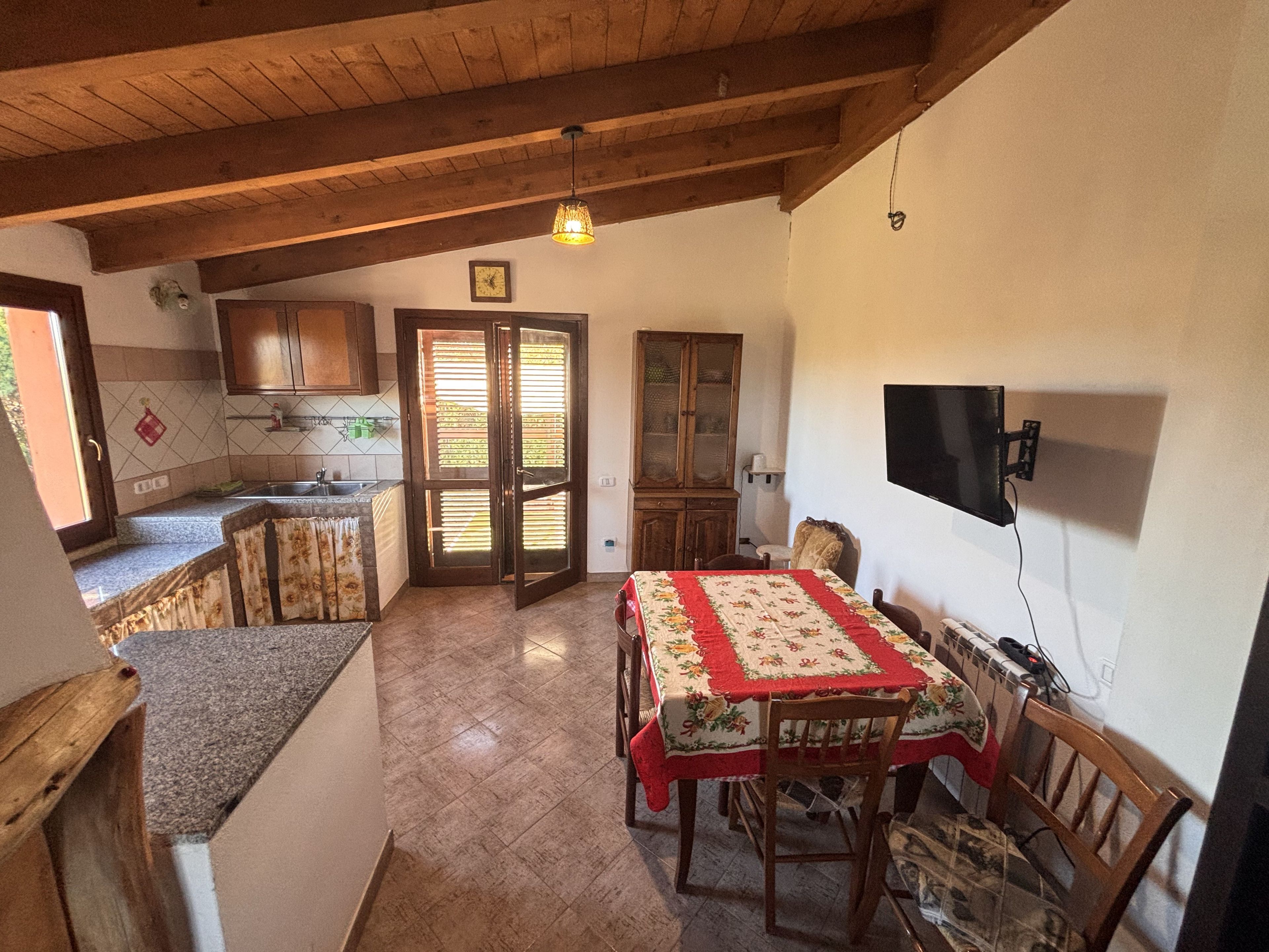 Una zona de menjador amb una taula, cadirees, un televisor fixat a la paret i una cuina amb armaris i un rentaplat.