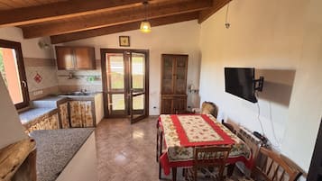 Una zona de menjador amb una taula, cadirees, un televisor fixat a la paret i una cuina amb armaris i un rentaplat.