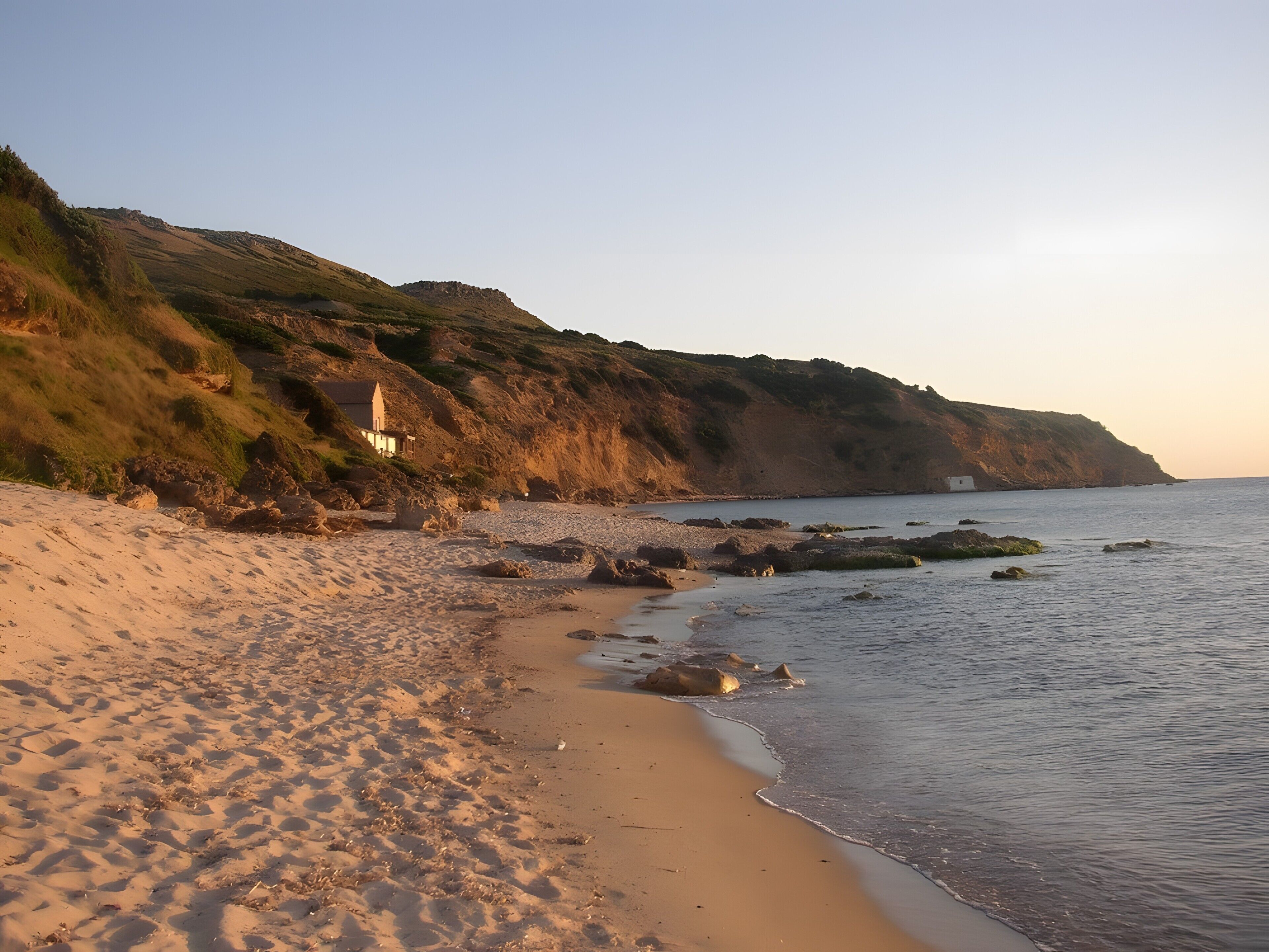 Una platja amb un edifici petit sobre un turó verd, costa rocosa i mar tranquil·la.