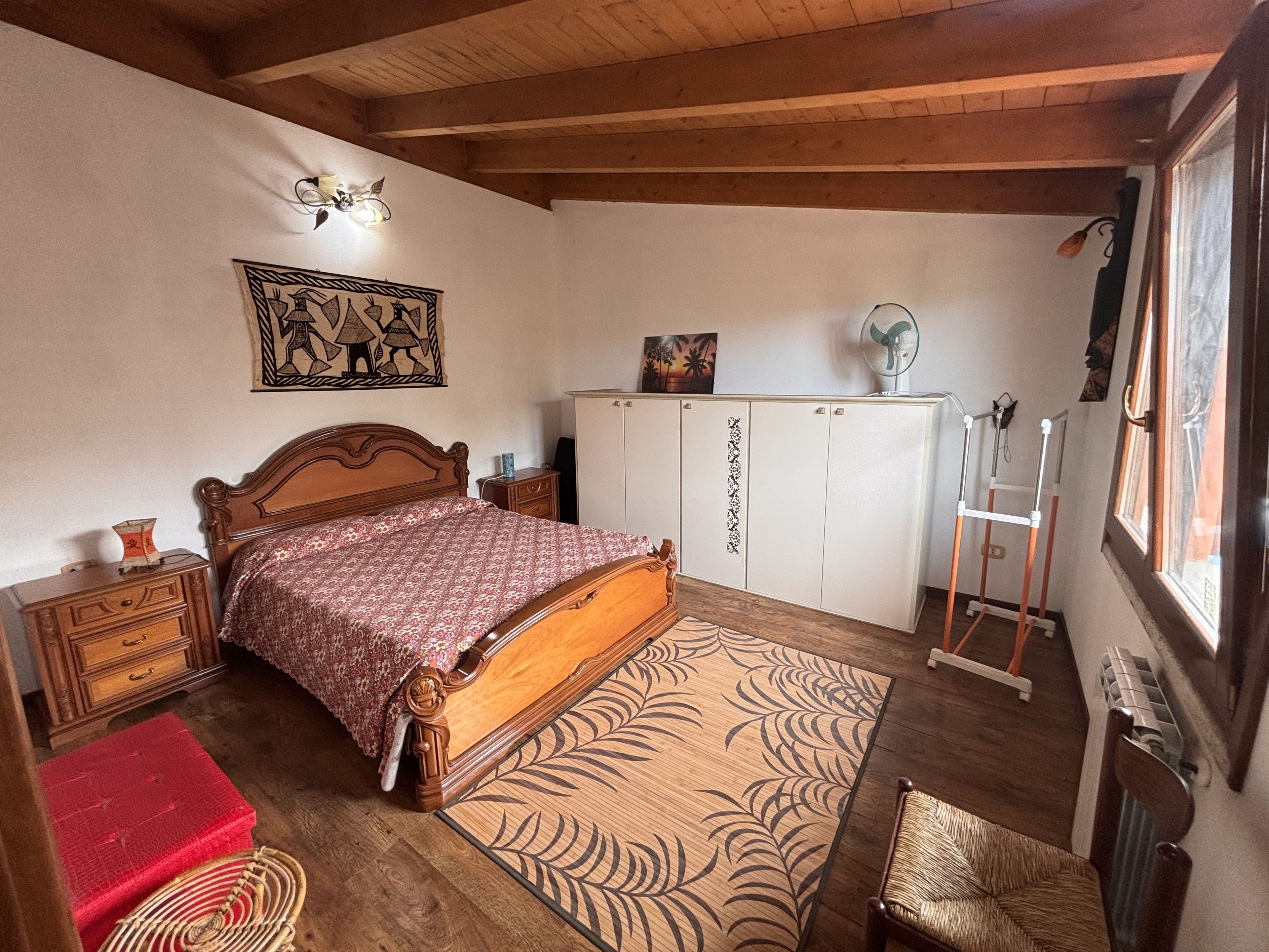 A bedroom with a wooden bed, a wardrobe, a bedside table, a red stool, a patterned rug, and a window with a view.