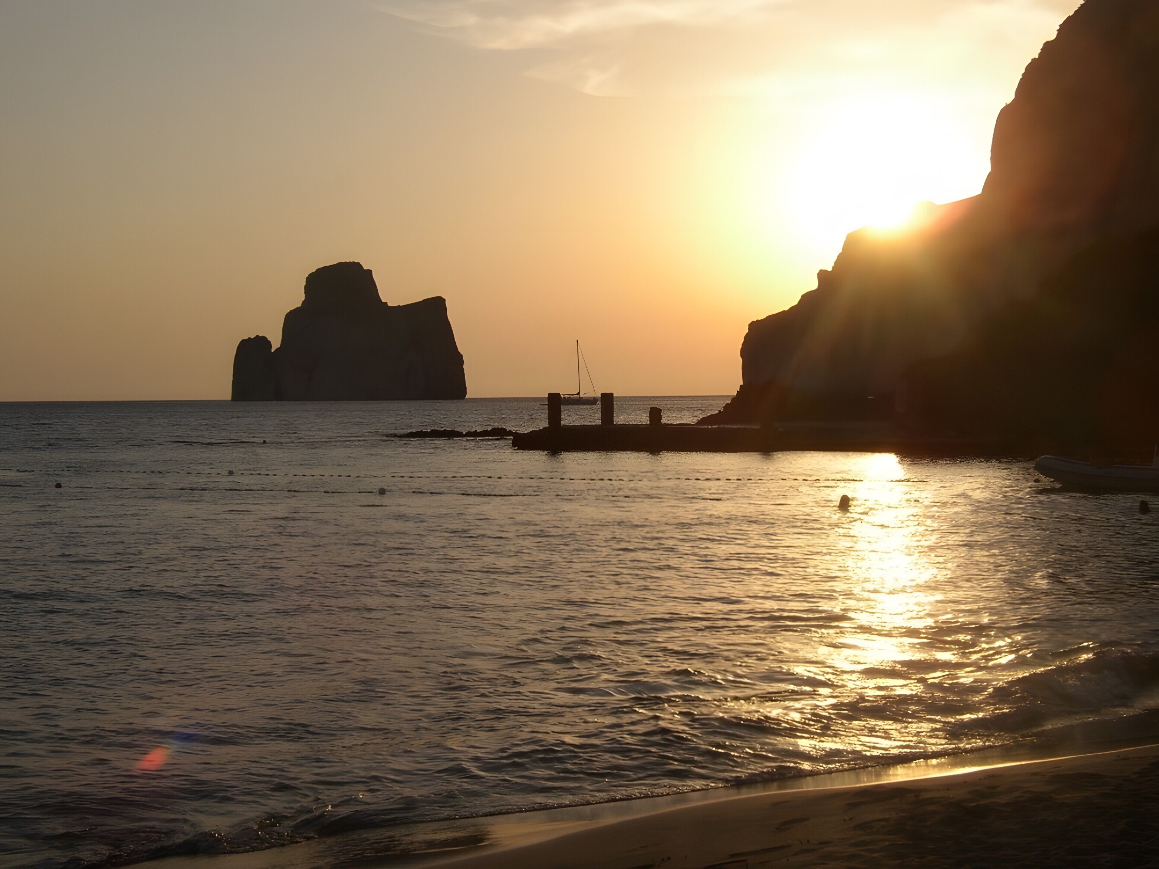A sunset over a calm sea with a rocky outcrop and a pier extending into the water.