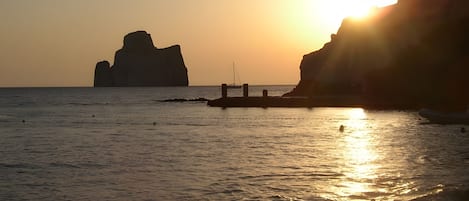A sunset over a calm sea with a rocky outcrop and a pier extending into the water.