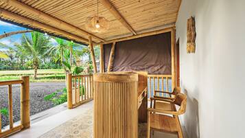 Un área de estar al aire libre con madera, que incluye una mesa y sillas, techo de bambú y vistas a un jardín con palmeras.