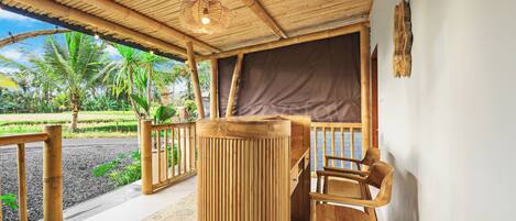 A wooden outdoor seating area with a table and chairs, a bamboo ceiling, and a view of a garden with palm trees.