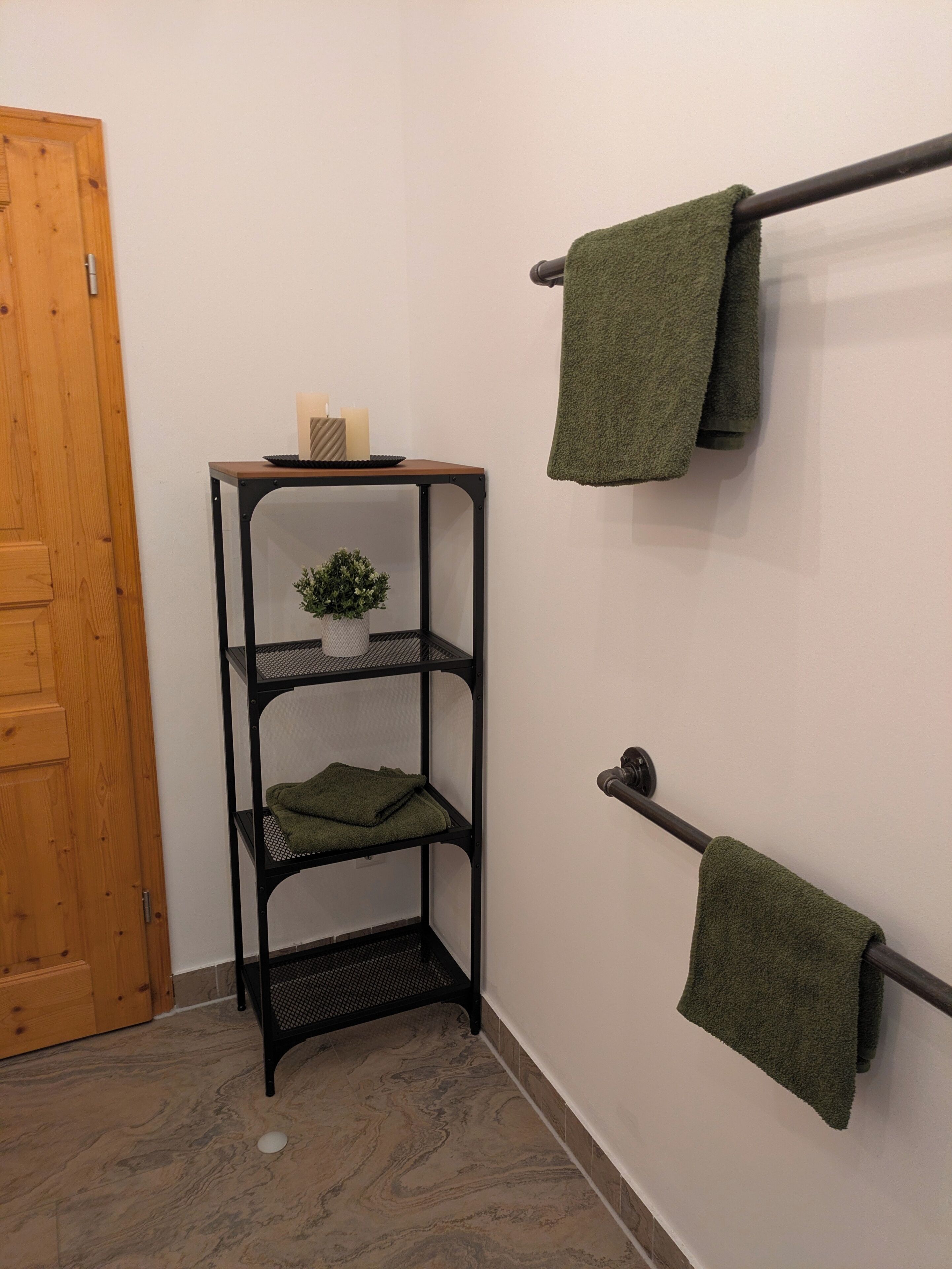 A corner with a wooden door, a black metal shelf with a plant, and two towels hanging on a towel rack.