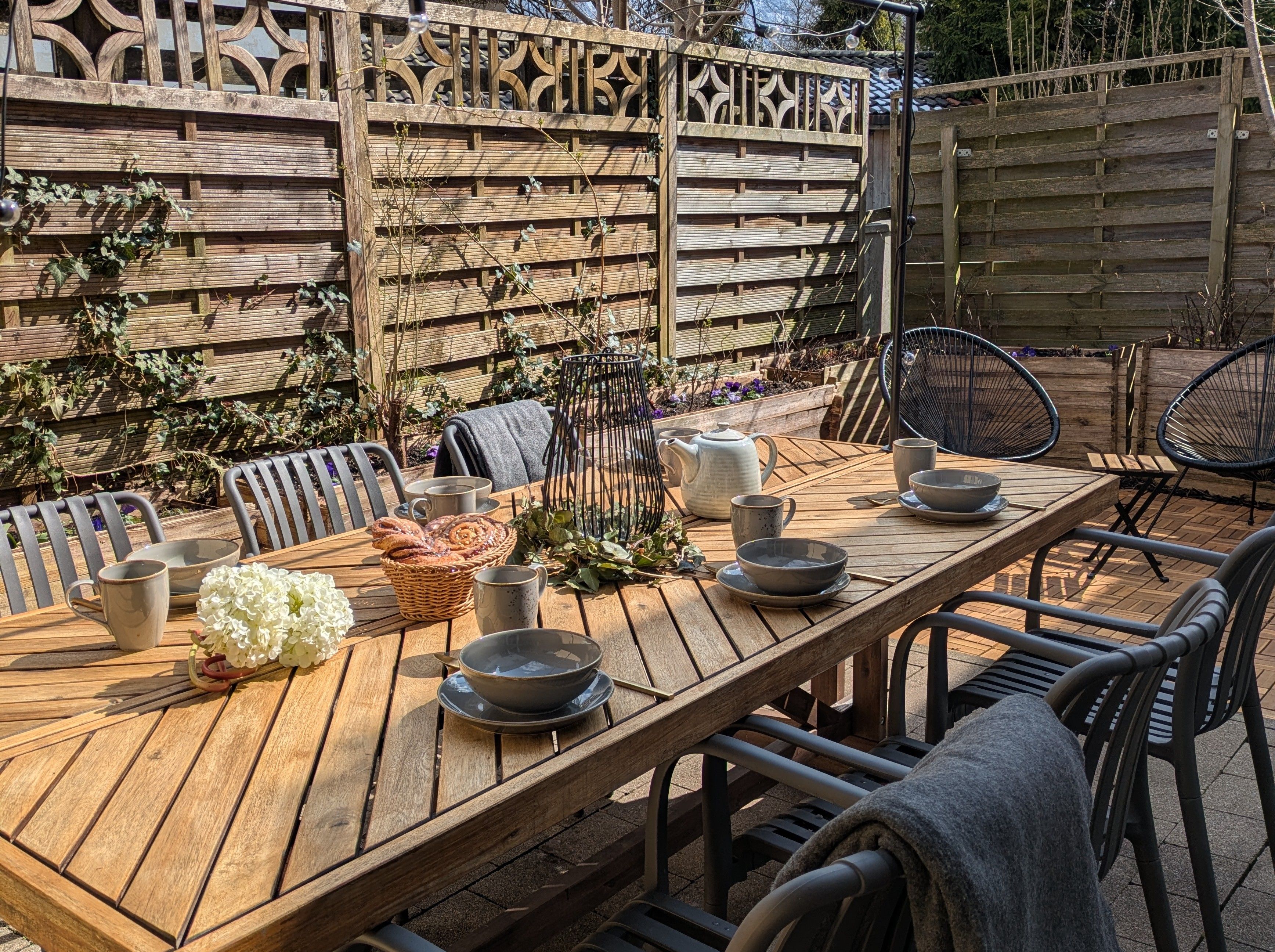 Una mesa de madera al aire libre con sillas, cuencos, tazas y una cesta de pan.