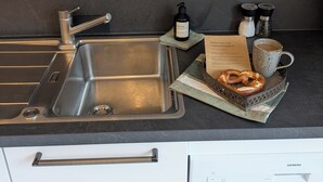 Eine Küche mit Spüle aus Edelstahl, einer Kaffeemaschine, einer Handseifenflasche, Salz- und Pfefferstreuern und einer Platte mit einem Brezel.
