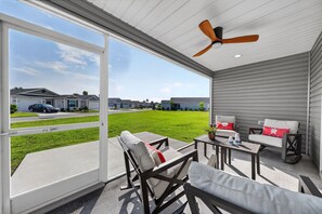 A covered outdoor area with a ceiling fan, a seating area with a table, and a view of a residential neighborhood.