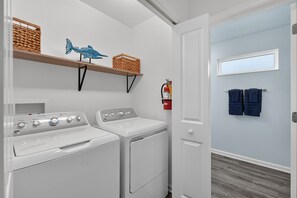 A laundry area with a washer and dryer, shelves with baskets, a fire extinguisher, and a door leading to a room with towels.