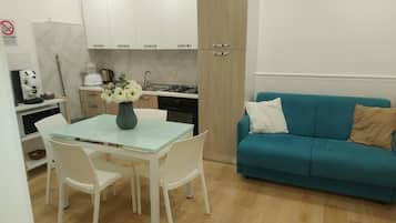 A compact kitchen with white cabinets, a dining table with chairs, and a teal sofa with cushions.