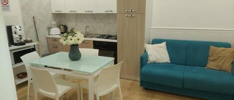 A compact kitchen with white cabinets, a dining table with chairs, and a teal sofa with cushions.