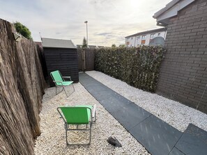 A backyard with two green chairs, a gravel path, a black fence, and a hedge. - Under 10m to Airport/Hospital/Hydro/Breahead/City (Renfrew)
