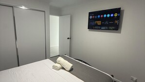 Ett hotellrum med en säng, en väggmonterad tv som visar olika appar, en stängd dörr och ett eluttag på väggen.