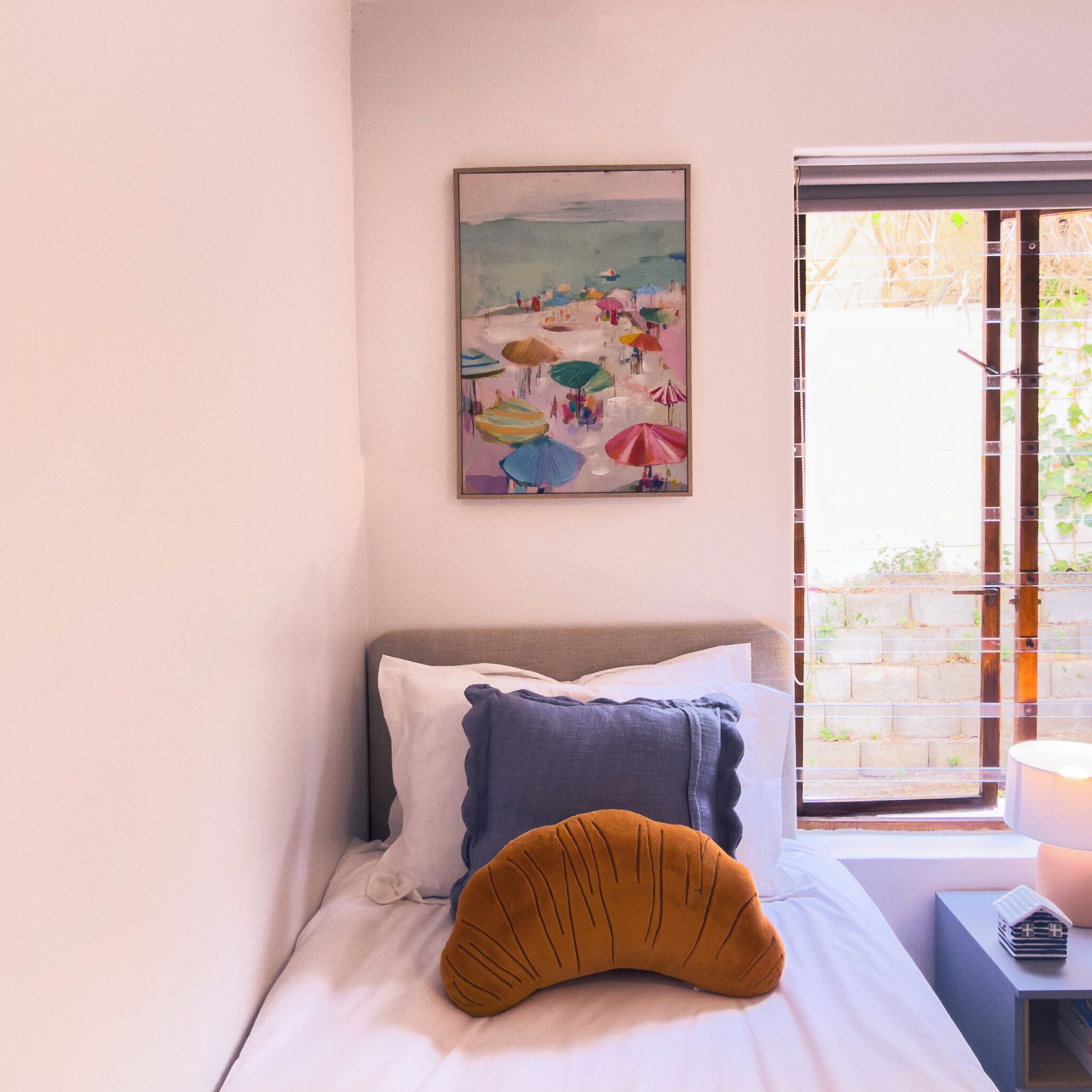 A bed with a pillow, a painting on the wall, and a window with a view of greenery.