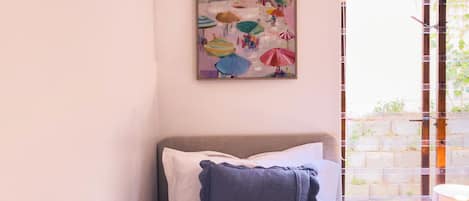A bed with a pillow, a painting on the wall, and a window with a view of greenery.