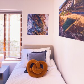 A bedroom with a bed, pillows, a unique orange cushion, a window with blinds, and two framed pictures on the wall. - Kalk. Beachfront Residence in Kalk Bay (Cape Town)