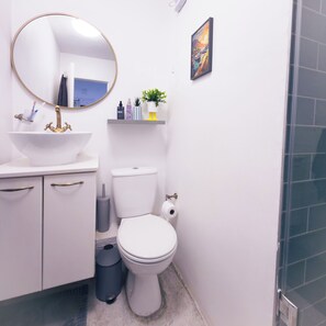 Kylpyhuone, jossa on valkoinen pesuallas, wc-istuin, roskakori, peili ja hylly, jossa on hygieniatuotteita.