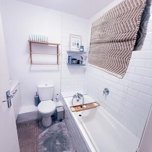 Ein Badezimmer mit Toilette, Waschbecken und Badewanne. Regale mit Pflegeprodukten und ein Handtuchhalter sind vorhanden.