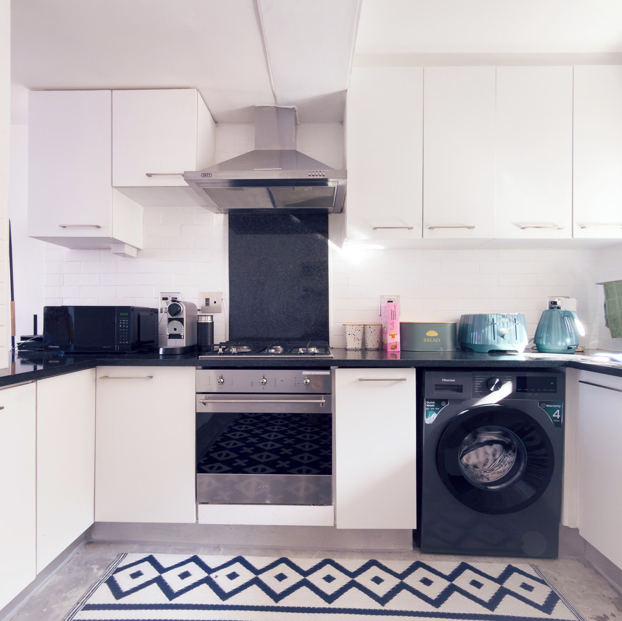 Une cuisine compacte avec des meubles blancs, une cuisinière noire, un four à micro-ondes, un lave-linge et un tapis à motifs.