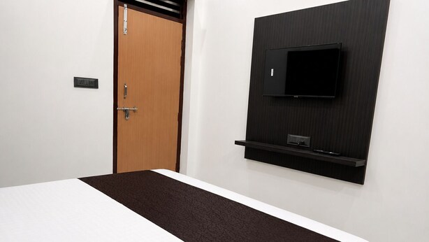 A hotel room with a bed, a wooden door, a TV mounted on the wall, and a small shelf below the TV. - Spacious 5-bedroom building in marvelous Mathura with AC (mathura)