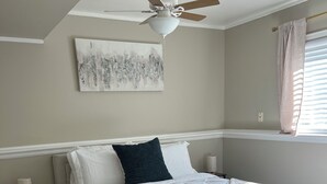 Un dormitorio con cama, ventilador de techo, ventana con cortinas y una obra de arte en la pared.