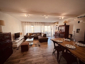 A living room with a sofa, a TV, a wooden dining table set for two, and a kitchen area with a refrigerator. - The Pink Hippopotamus – Duplex with a terrace overlooking the M (Sète)