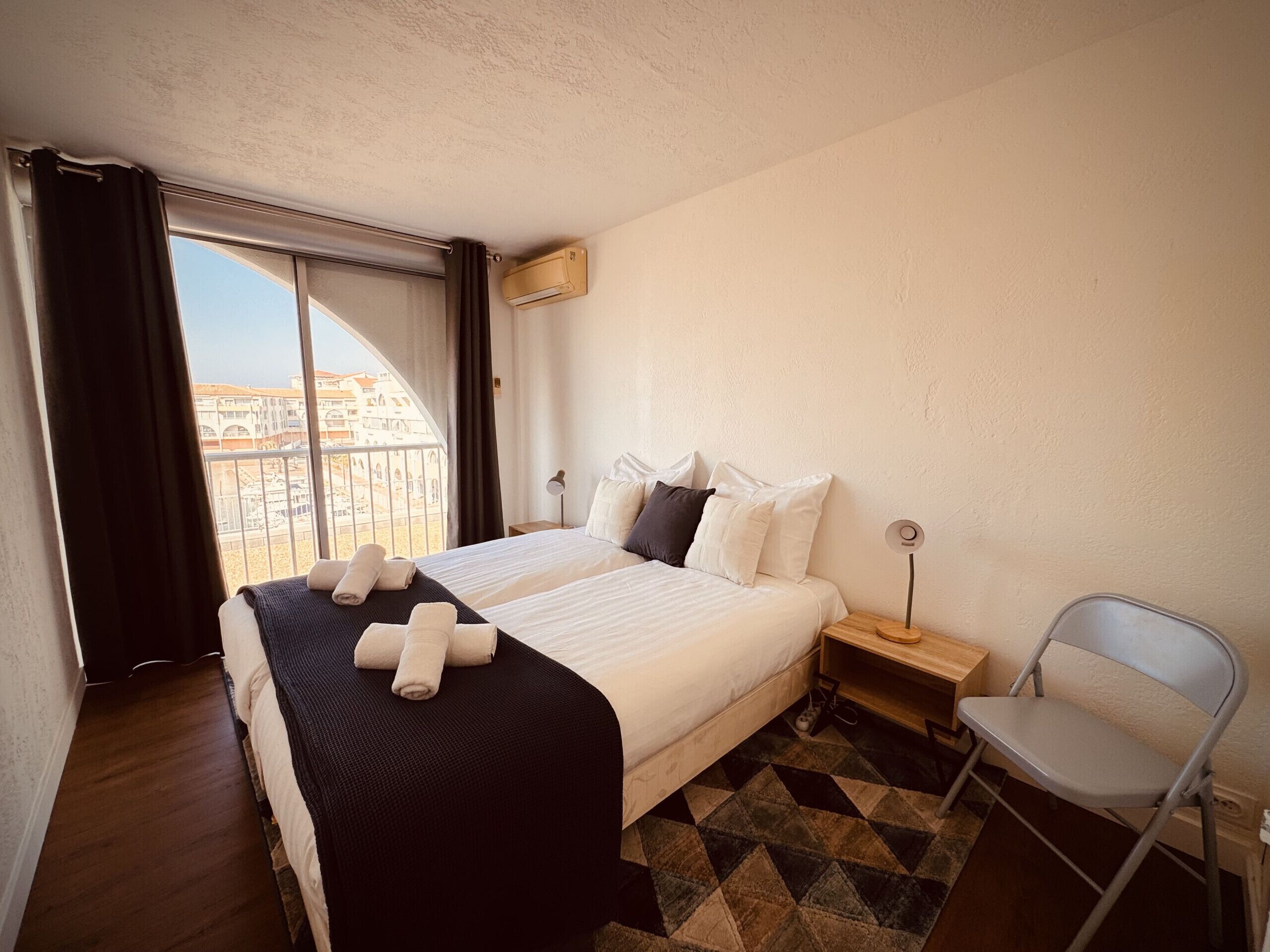 A hotel room with a large bed, a chair, a nightstand, and a view of buildings through a window.