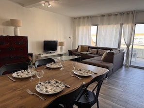 Een eethoek met een houten tafel gedekt voor twee personen, een televisie op een standaard, een bank en uitzicht op het buitenlands gebied door grote ramen.