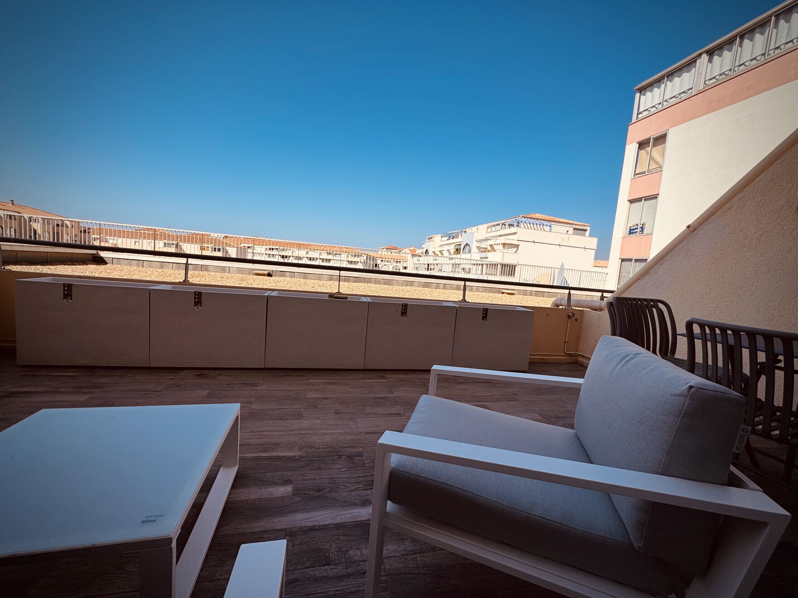 A rooftop terrace with a white table and chair, a cushioned sofa, and a view of buildings in the distance.