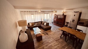 A modern living room with a sectional sofa, a dining table with chairs, a kitchen area with a microwave and refrigerator, and a large window with curtains. - The Pink Hippopotamus – Duplex with a terrace overlooking the M (Sète)