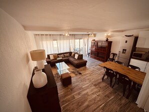 A modern living room with a sectional sofa, a dining table with chairs, a kitchen area with a microwave and refrigerator, and a large window with curtains. - The Pink Hippopotamus – Duplex with a terrace overlooking the M (Sète)