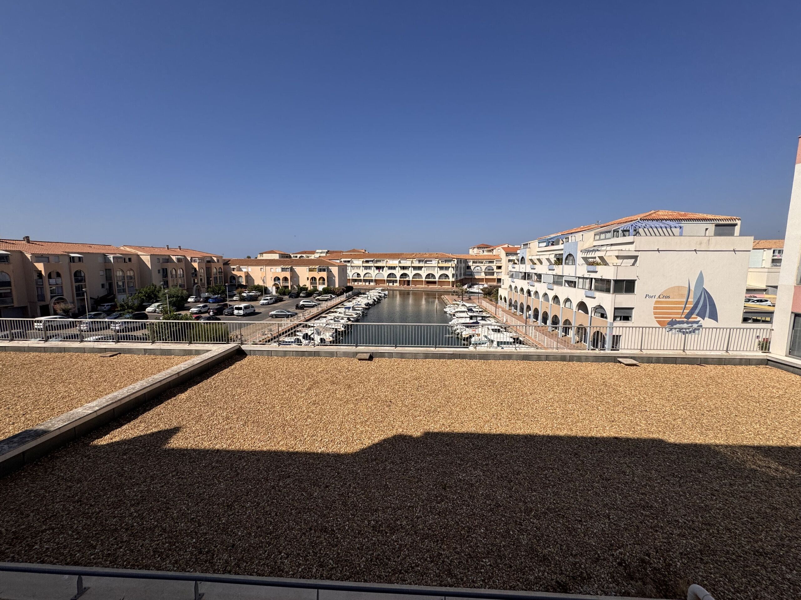 A marina with boats docked, a building with a sailboat logo, and a clear sky.