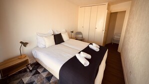 A neatly made bed with white linens and a black throw blanket, a bedside lamp, and a chair in a room with a tiled floor and a wardrobe. - The Pink Hippopotamus – Duplex with a terrace overlooking the M (Sète)