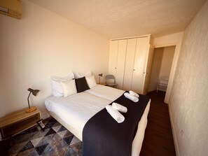 A neatly made bed with white linens and a black throw blanket, a bedside lamp, and a chair in a room with a tiled floor and a wardrobe. - The Pink Hippopotamus – Duplex with a terrace overlooking the M (Sète)
