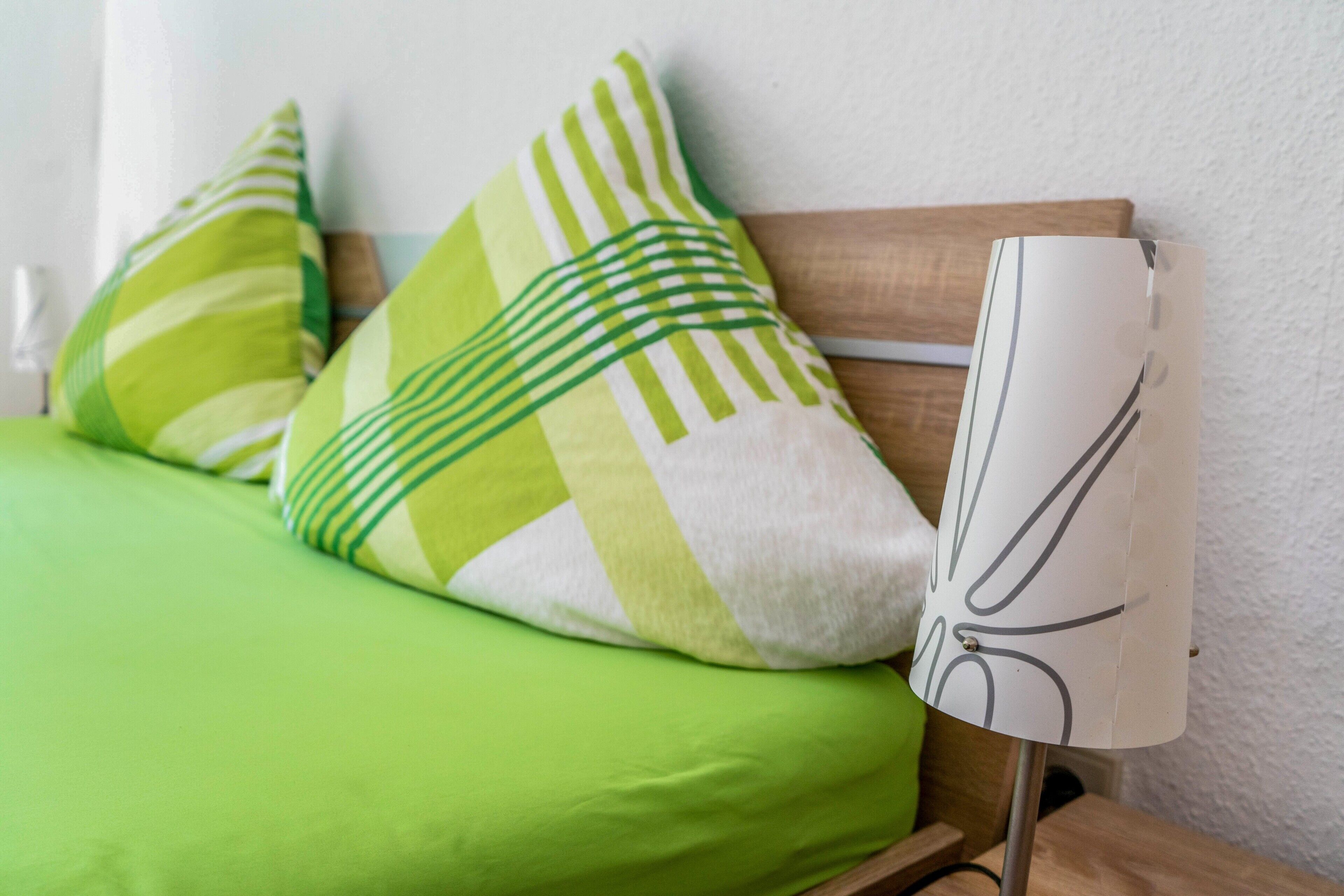 Ein Bett mit grüner Bettdecke und gestreiften Kissen, eine schlichte, weiße Lampe und ein Holz-Kopfschild.