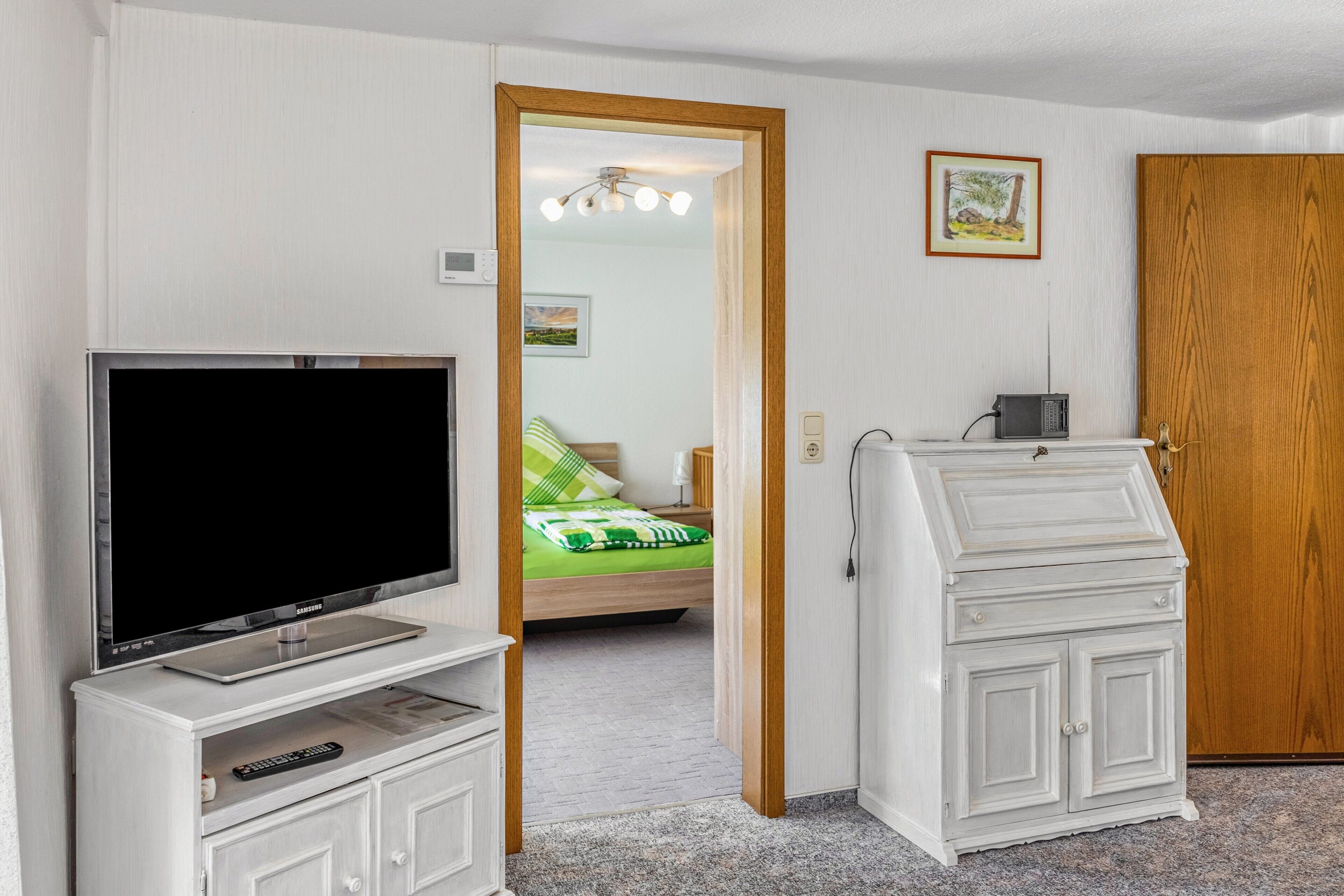 Ein Hotelzimmer mit Fernseher, einem kleinen Schrank, einem Bett mit einer grün-weiß gestreiften Decke, einer Holztür und einem kleinen Tisch mit einem Radio.